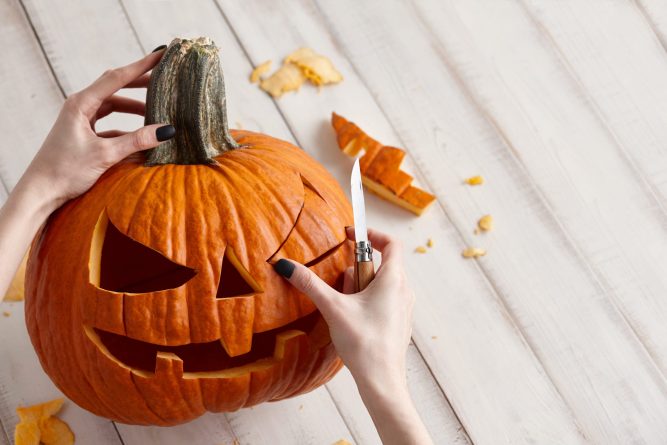 Woman carving big orange pumpkin into jack-o-lantern for Halloween holiday decoration on white wooden planks, close up view