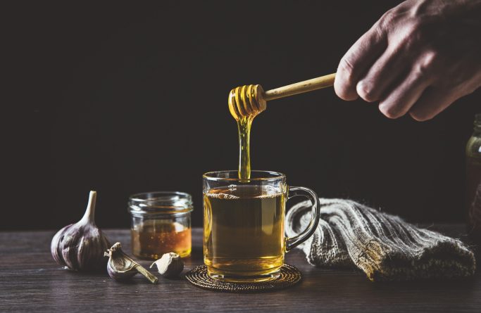 Man hand holding wooden honey dipper, honey spoon on top of glass of tea/ medicine and dripping honey in hot tea. Knitted socks, small jar of honey, garlic on wooden table against black background.