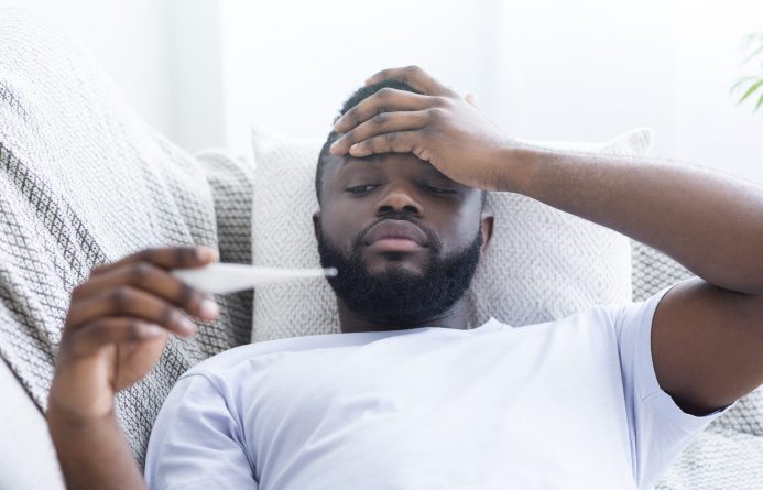 Must be sick. Sad african-american guy with fever measuring his body temperature, lying on couch at home